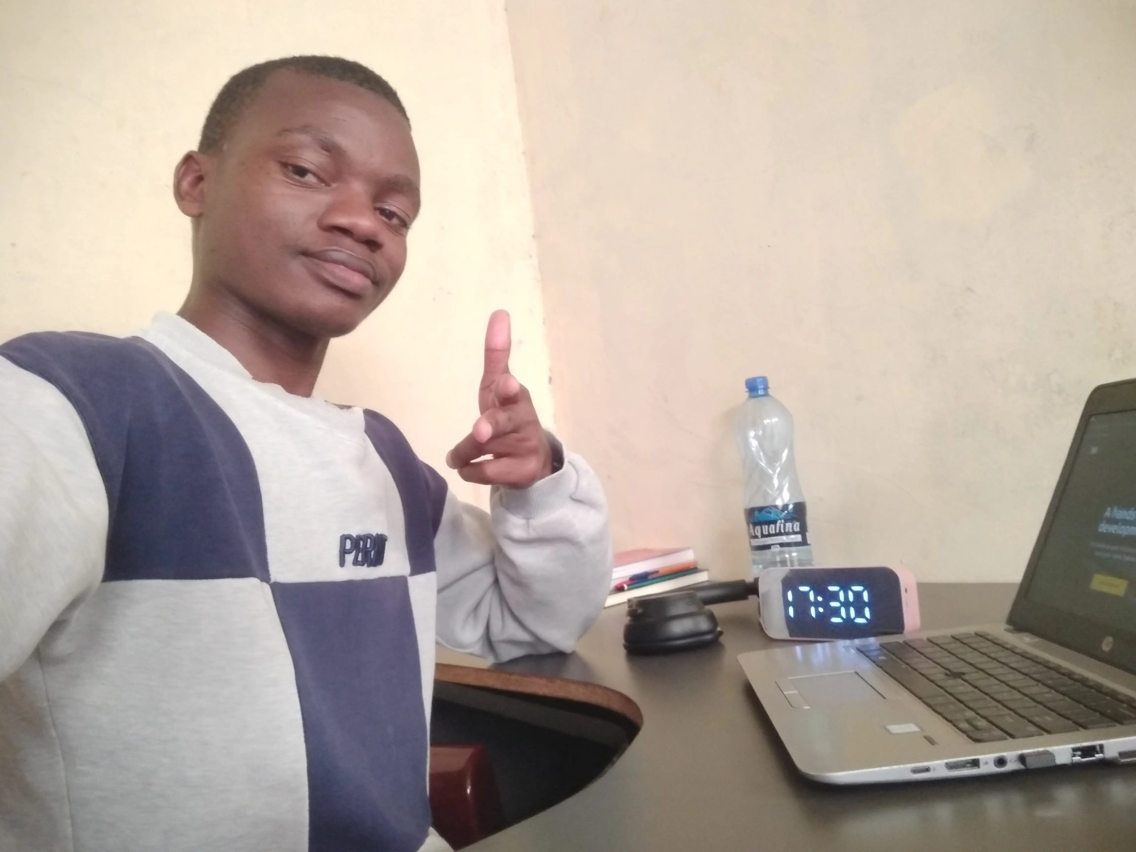 Jarvis Wuod in a blue and white striped sweater sits at a wooden desk, pointing at the camera with a slight smile. On the desk are a laptop computer, a digital clock displaying 1:30